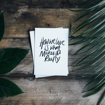 A piece of paper that says "how we live is what makes us real" on a wooden surface with leaves on both sides