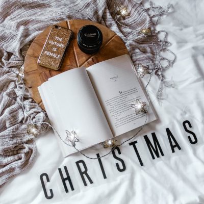 Flatlay of someone's bed with an open book, some lights, a canon lens, a cellphone case that says "The Futer is Female" and words that say "Christmas"